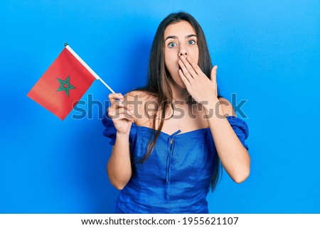 Young brunette teenager holding morocco flag covering mouth with hand, shocked and afraid for mistake. surprised expression 