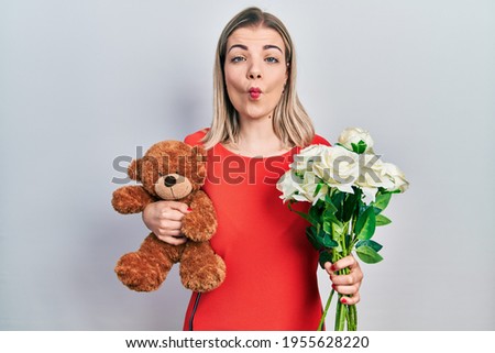 Beautiful caucasian woman holding bear and bouquet of flowers for anniversary making fish face with mouth and squinting eyes, crazy and comical. 