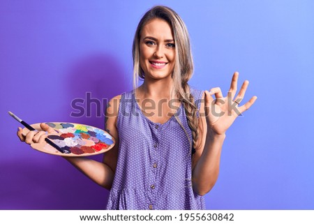 Young beautiful blonde artist woman drawing using paintbrush and palette with colors doing ok sign with fingers, smiling friendly gesturing excellent symbol