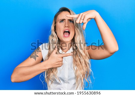 Beautiful young blonde woman doing picture frame gesture with hands angry and mad screaming frustrated and furious, shouting with anger looking up. 