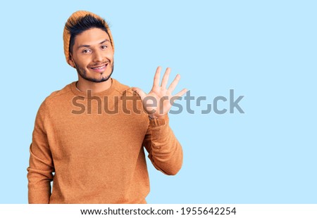 Handsome latin american young man wearing winter sweater and wool hat showing and pointing up with fingers number five while smiling confident and happy. 
