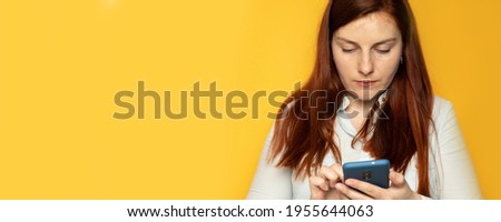 Cheerful young beautiful woman surfing smartphone on a yellow plain background wall in workplace