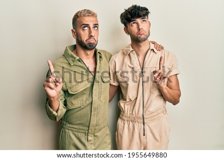 Homosexual gay couple standing together wearing casual jumpsuit pointing up looking sad and upset, indicating direction with fingers, unhappy and depressed. 