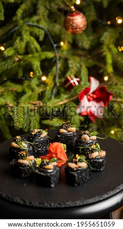Sushi with black rice, crab meat, avocado, smoked salmon mousse, oar caviar, masago, shrimp cocktail and edible gold leaf with ginger on black table for Christmas with a Christmas tree on background.