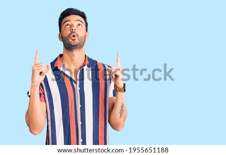 Young handsome hispanic man wearing casual  summer shirt amazed and surprised looking up and pointing with fingers and raised arms.  Royalty-Free Stock Photo #1955651188
