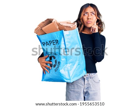 Young beautiful mixed race woman holding recycling wastebasket with paper and cardboard serious face thinking about question with hand on chin, thoughtful about confusing idea 