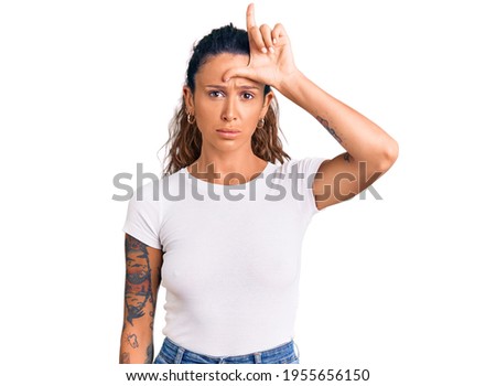Young hispanic woman with tattoo wearing casual white tshirt making fun of people with fingers on forehead doing loser gesture mocking and insulting. 