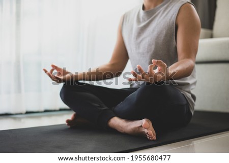 Man in a gray sleeveless shirt and black pants sat on the exercise cushions, practicing meditation, sitting in a lotus pose alone at home. During the quarantine due to the spread of the coronavirus. Royalty-Free Stock Photo #1955680477