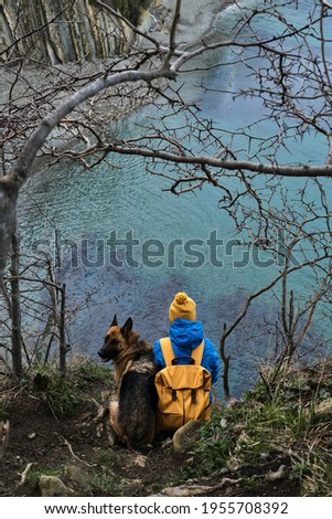 Dog with owner on the sea. Girl with yellow backpack and hat sits on the edge of cliff overlooking the blue ocean next to black and red German shepherd friend. Walking and traveling with dog.