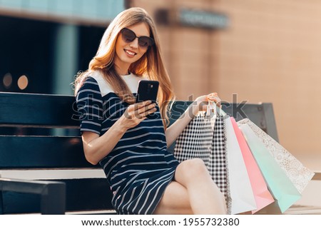 attractive happy beautiful young woman in sunglasses is sitting on a bench near a shopping center with shopping bags, taking a selfie on her mobile phone. Shopping, fashion, online shopping