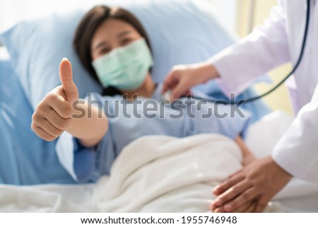 Happy Asian female patient wears a mask, lies on the bed, and raises her thumb up. When a doctor uses a stethoscope to listen to the lungs. Concept of believe in treatment And insurance coverage Royalty-Free Stock Photo #1955746948