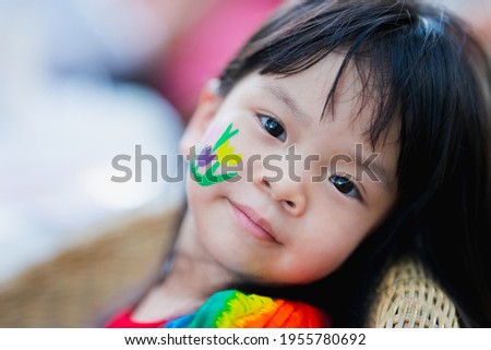 Closeup of cute Asian girl face sitting on chair made of wood. Watercolors are cartoon flower on skin of kid. Happy child looks at camera and smiling. Children are excited Easter day. Baby 4 years old