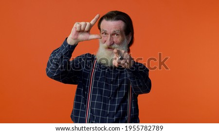 Elderly bearded gray-haired man showing loser gesture and pointing on you, blaming accusing for unsuccess, expressing disrespect, mocking your failure. Senior old grandfather on orange background