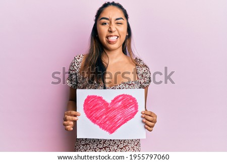 Young latin woman holding heart draw sticking tongue out happy with funny expression. 