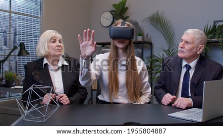 Young woman wearing virtual reality glasses tries 3D app for VR helmet while company colleagues supporting her in modern office. Workers team using innovative future technology through VR headset