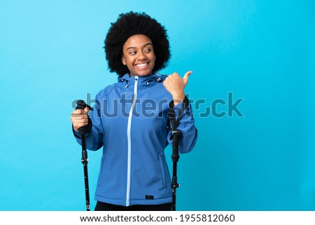 Young Africa American with backpack and trekking poles isolated on blue background pointing to the side to present a product