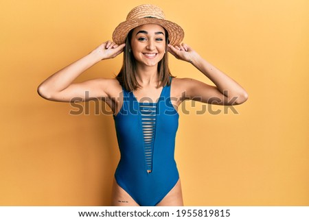 Young brunette girl wearing swimsuit and summer hat smiling pulling ears with fingers, funny gesture. audition problem 