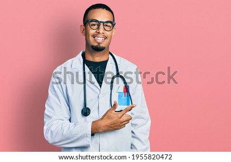 Young african american man wearing doctor uniform and stethoscope cheerful with a smile on face pointing with hand and finger up to the side with happy and natural expression 