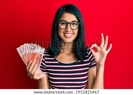 Beautiful asian young woman holding 100 norwegian krone banknotes doing ok sign with fingers, smiling friendly gesturing excellent symbol 