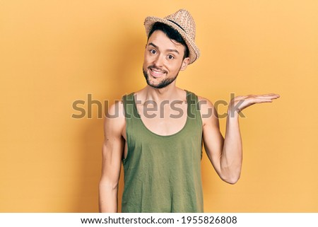 Young hispanic man wearing summer hat smiling cheerful presenting and pointing with palm of hand looking at the camera. 