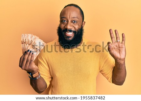 Young african american man holding south african 20 rand banknotes doing ok sign with fingers, smiling friendly gesturing excellent symbol 