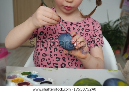 Adorable baby girl 4-5 years old in a pink dress paints Easter eggs with watercolors for Easter and play. The child is engaged in creativity and prepares for Easter.
