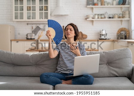 Tired millennial woman suffers from stuffiness and an inoperative air conditioner, waving blue fan sitting on couch at home working on laptop computer. Overheating high temperature, hot summer weather Royalty-Free Stock Photo #1955951185