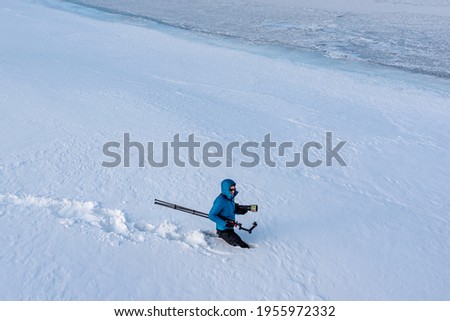 A man, photographer walking across a winter landscape with very deep snow with camera, tripod and a blue jacket with white background. 