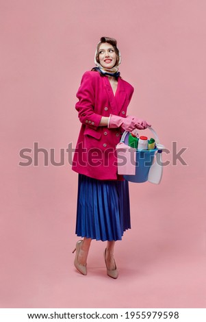 A happy woman holds a bucket of cleaning tools in her hands. Bright makeup. Pin-up style. Pink background. Cleaning concept.