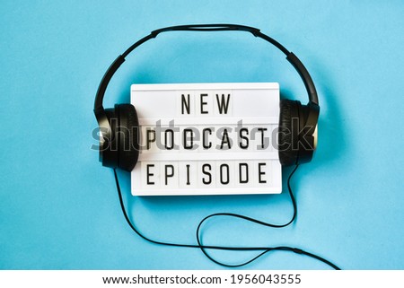 Top view of words NEW PODCAST EPISODE on light box on blue background with headphones. Podcasting concept.  Royalty-Free Stock Photo #1956043555