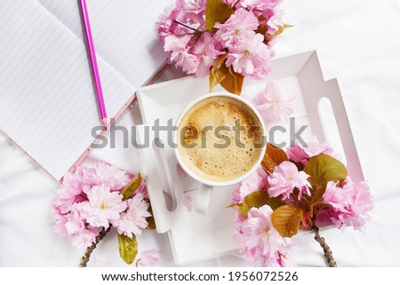 Big cup of coffee with milk and notebook on the white bedsheet. Sakura flowers around cut of hot coffee. Top view. Spring time. Wooden plate with coffee and flowers. Work from home or anywhere. Cozy.
