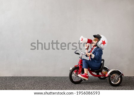 Happy child wearing suit riding bicycle against grey concrete wall background. Funny kid shouting through loudspeaker. Childhood dreams and business idea concept Royalty-Free Stock Photo #1956086593