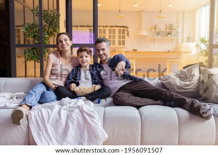 Smiling laughing parent showing funny video to son on television. Parent and son watching comedy movie on tv-set at home. Happy family sitting on comfort sofa. Cozy evening in living room interior
