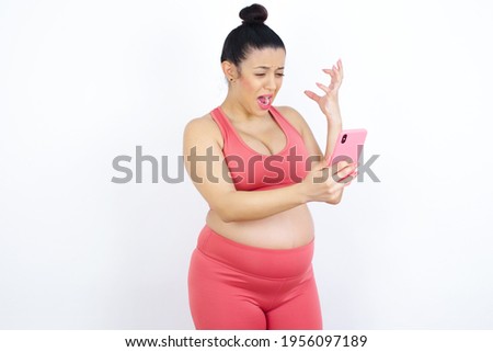 Photo of outraged annoyed young beautiful Arab pregnant woman in sports clothes against white wall holds cell phone, makes call, argues with colleague,  expresses negative emotions. People and anger.