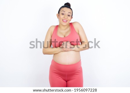 Happy young beautiful Arab pregnant woman in sports clothes against white wall stands against orange studio wall keeps hands on heart, swears be loyal, expresses gratitude. Honesty concept.