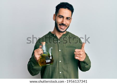 Young hispanic man holding olive oil can smiling happy and positive, thumb up doing excellent and approval sign 