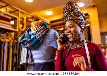 tanzanian woman with snake print turban over hear working in fabrics shop calling to client by smartphone Royalty-Free Stock Photo #1956159304