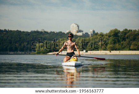 Handsome athletic man with muscular body swims on sup board on pond on summer hot day, wears sunglasses
