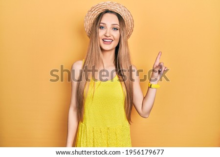 Young blonde girl wearing summer hat with a big smile on face, pointing with hand finger to the side looking at the camera. 