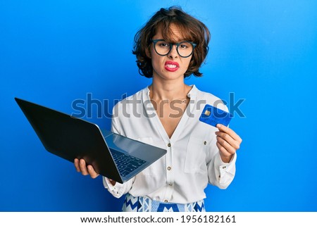 Young brunette woman wearing business style holding laptop and credit card clueless and confused expression. doubt concept. 