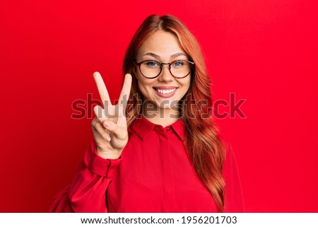 Young beautiful redhead woman wearing casual clothes and glasses over red background showing and pointing up with fingers number two while smiling confident and happy. 