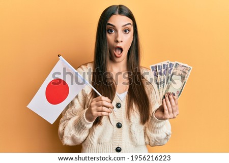 Beautiful brunette young woman holding japan flag and yen banknotes afraid and shocked with surprise and amazed expression, fear and excited face. 