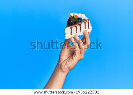 Hand of hispanic man holding slice of chocolate cake over isolated blue background. Royalty-Free Stock Photo #1956222301