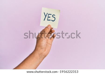 Hand of hispanic man holding yes reminder paper over isolated pink background.