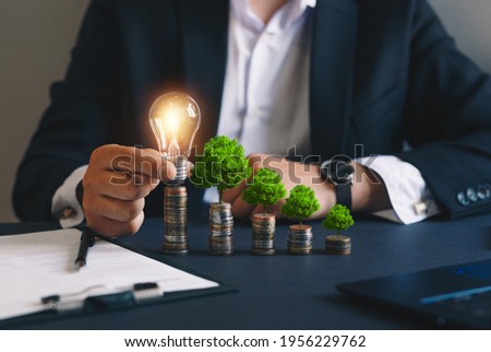 Businessman holding a light bulb and stacks of money growing with green trees. Concept of
pass and increase of renewable energy. Alternative sources of energy. Green energy, eco energy. 