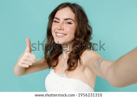 Close up of smiling bride young woman in white wedding dress doing selfie shot on mobile phone showing thumb up like gesture isolated on blue turquoise background. Ceremony celebration party concept