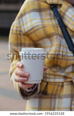 A cup of coffee in a disposable cup in the hands of a stylish woman in the city. Cup design mockup