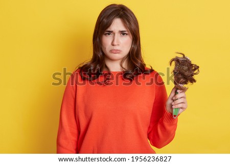 Sorrowful girl with purse lips showing to camera comb with her lost hair, has problem, needs treatment, expressing negative emotions, being upset, isolated over yellow background.