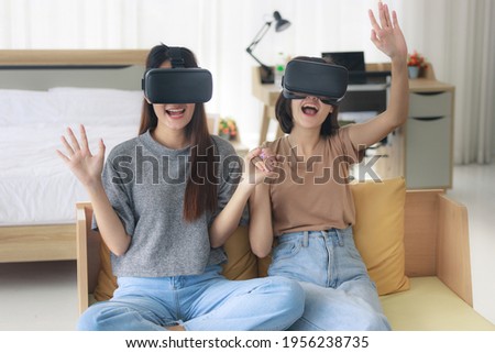 Two Asian teenage girls are Intimate friends with short and long hair wearing VR glasses for play games and watching a movie or entertainment in the bedroom. Young women are enjoying leisure activitie