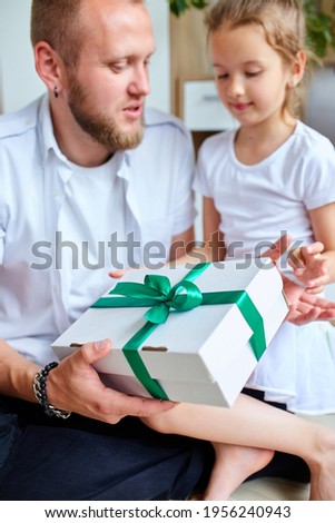 Little girl is giving her handsome father a gift box on Father's day, smiling daughter congratulating dad and giving present on birthday at home. I love you, dad. Happy Father's Day.